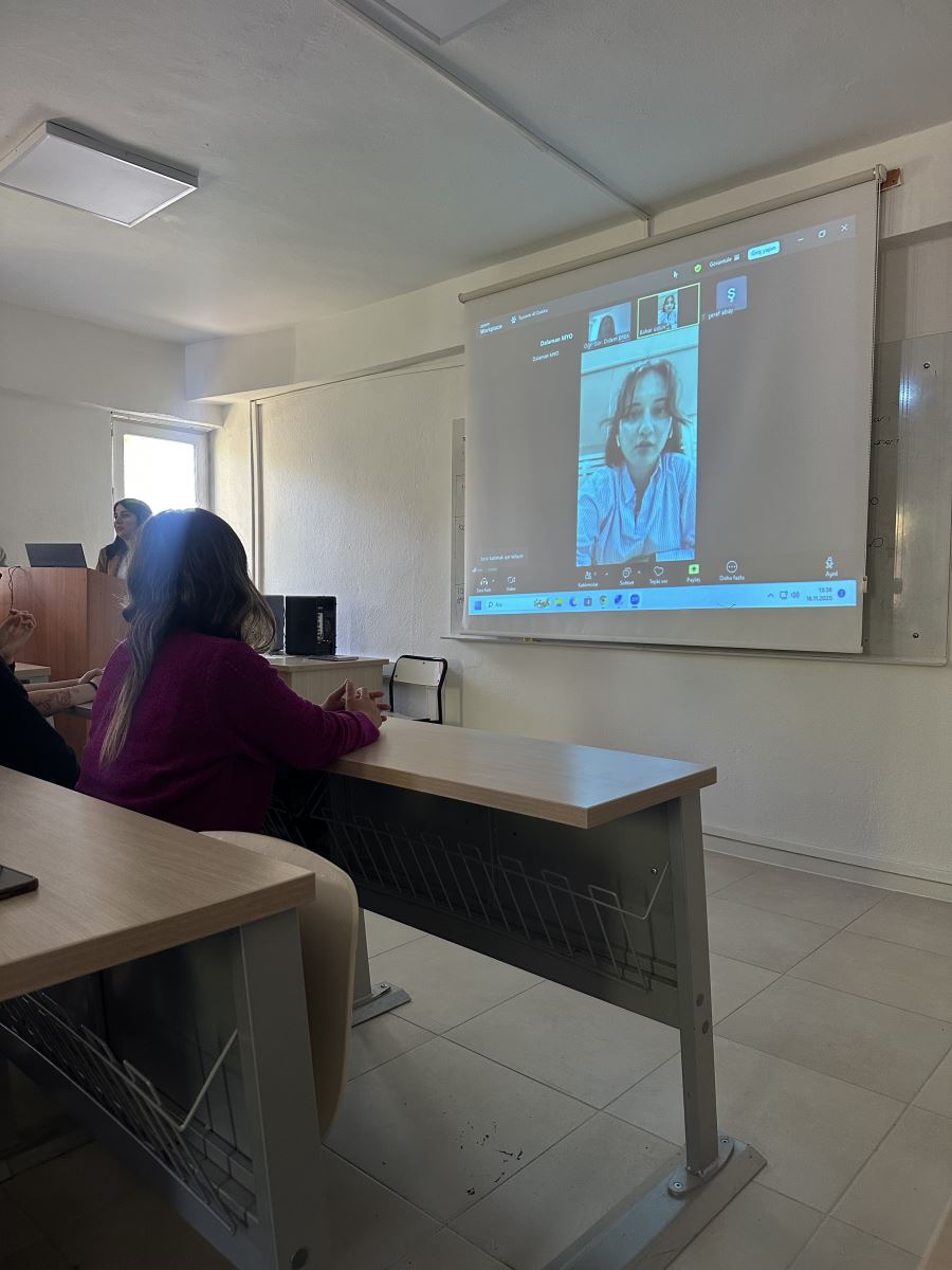A classroom is shown with students seated at desks. At the front of the room, a projection screen displays an online meeting. On the screen, a woman wearing a blue shirt and earphones is speaking to the students virtually. In the front row, a student in a pink sweater is watching the screen attentively. On the left side, an instructor is standing behind a podium, working on a laptop. The room is bright with white walls, and the atmosphere is calm as the students follow the presentation.
